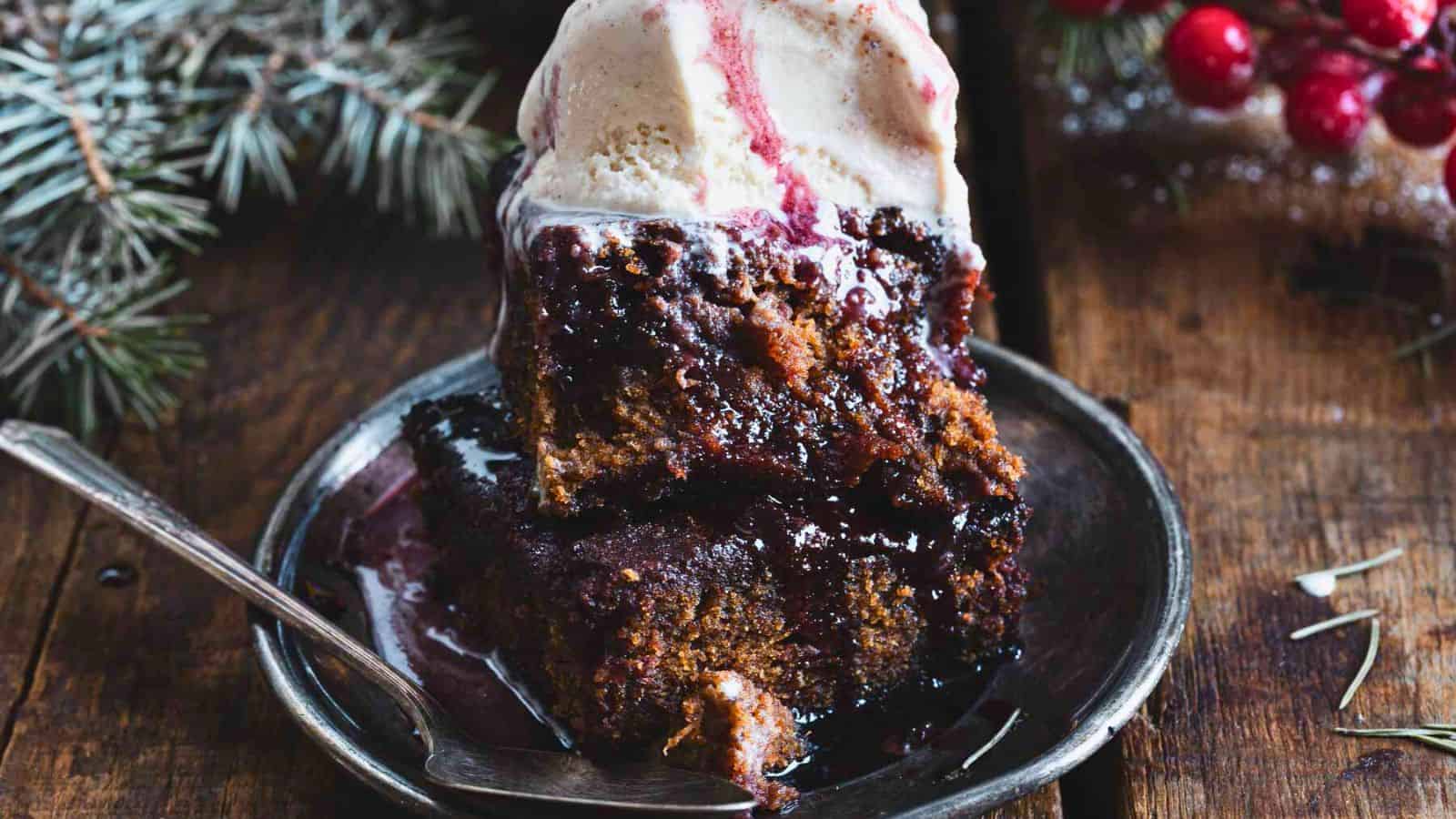 A slice of dark cake with a scoop of vanilla ice cream on top, drizzled with berry sauce, sits on a plate. Red berries and evergreen branches are in the background on a wooden surface.