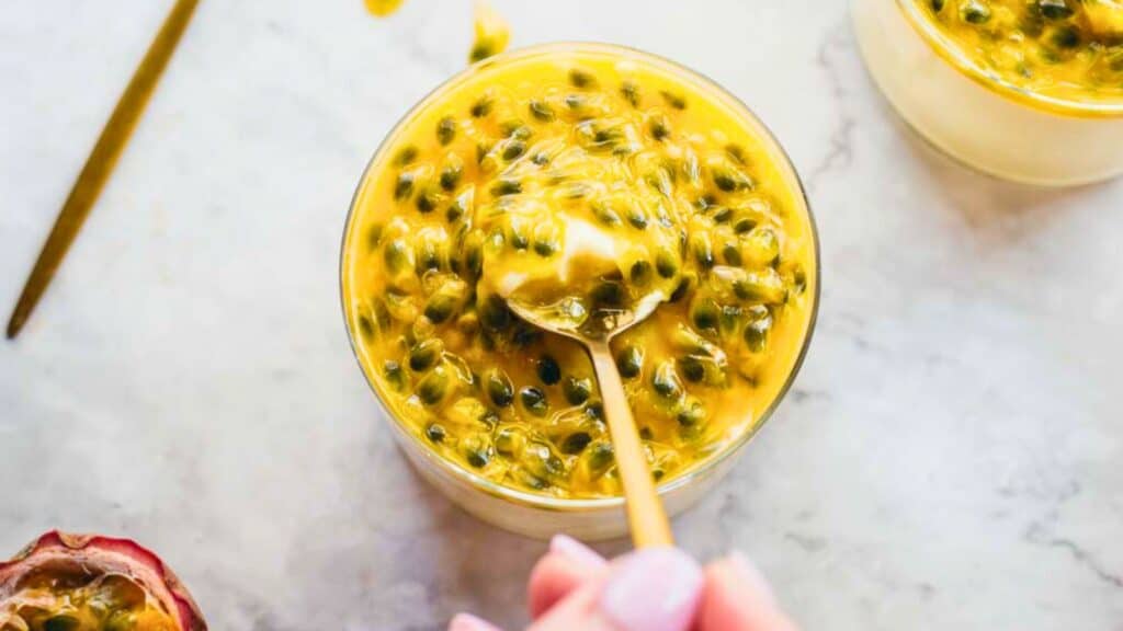 A hand holding a spoon scoops passion fruit pulp from a glass container on a marble surface.