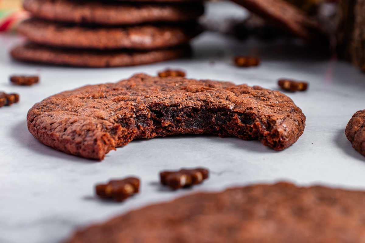 A chocolate cookie with a bite taken out. Several whole cookies and small chocolate pieces are in the background on a white surface.