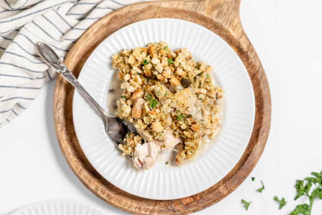 A plate with chicken and stuffing, topped with herbs, on a wooden serving board. A spoon rests on the plate. Striped cloth nearby.