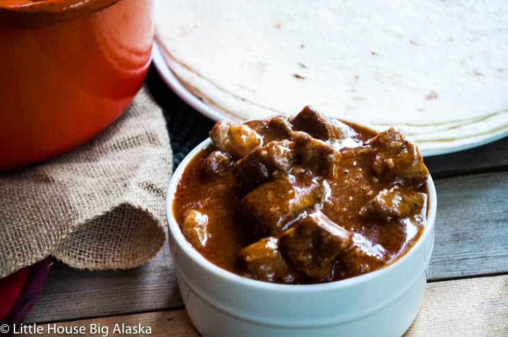 A bowl of beef stew in rich sauce beside a stack of tortillas, with an orange pot partially visible.