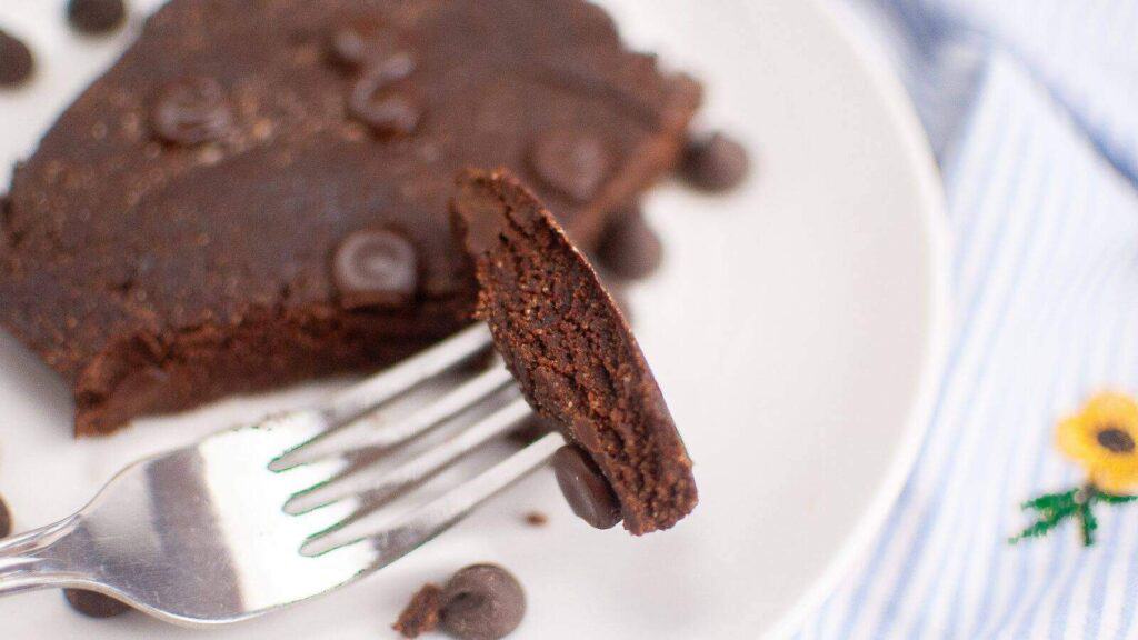 A piece of chocolate brownie with chocolate chips is held on a fork above a white plate. Additional chocolate chips are scattered around the plate.