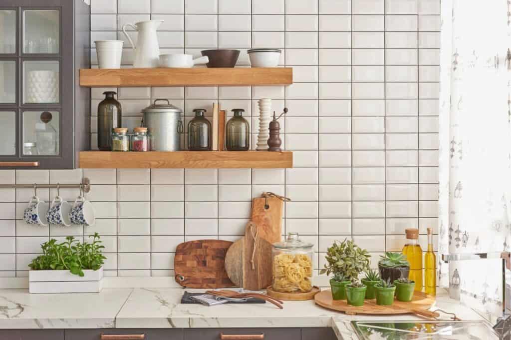 A freshly refreshed kitchen counter features herbs, cutting boards, olive oil, and pasta in a jar nestled among potted plants. Wooden shelves display jars, bowls, and spices against a white tiled backsplash, while sunlight filters softly through a sheer curtain.