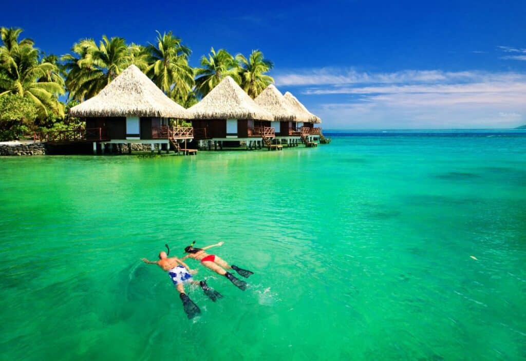 Two people snorkel in clear turquoise water near thatched-roof overwater bungalows, with palm trees and a blue sky in the background.