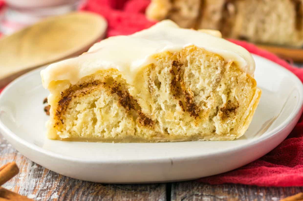 A close-up image of a slice of Cracker Barrel Cinnamon Roll Pie, showing soft, swirled cinnamon roll layers inside a flaky pie crust, topped with a thick, creamy white icing, served on a white plate.