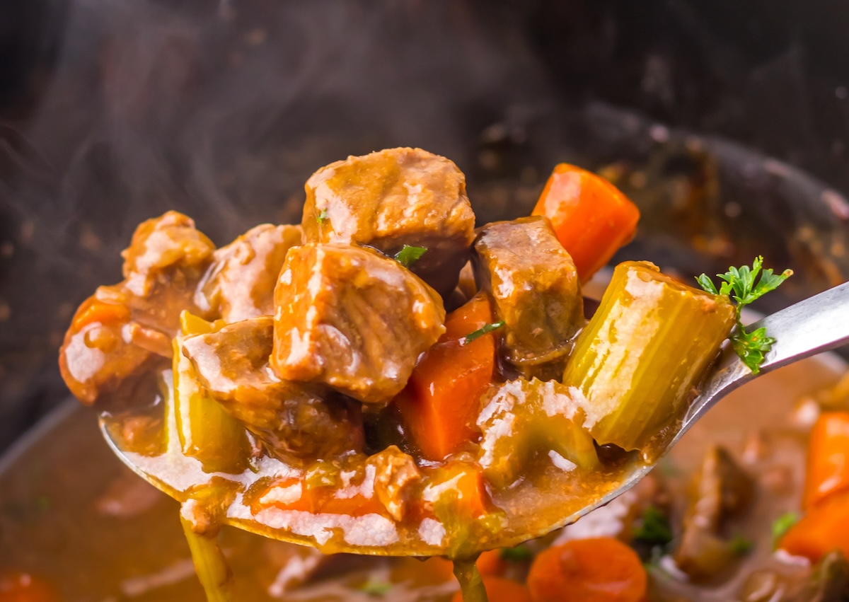 A spoonful of beef stew with carrots, celery, and parsley is being lifted from a pot. Steam is visible rising from the stew.