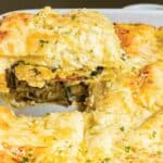 A slice of cheesy vegetable lasagna being lifted from a white baking dish, showing layers of pasta, spinach, and melted cheese.