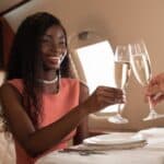 A woman in a pink dress clinks champagne glasses with another person inside a private jet.