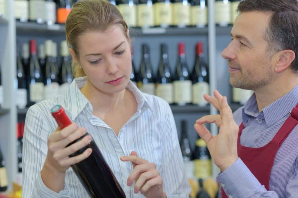 A woman examines a wine bottle while a man in an apron gestures at a wine shop.