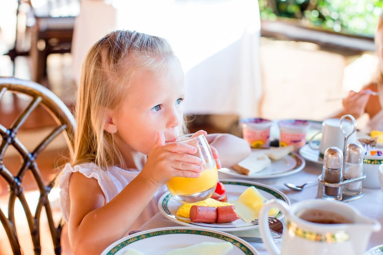 A young girl drinks orange juice at a table set with breakfast items, including eggs, sausage, and fruit.