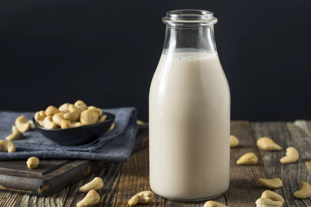 A glass bottle of cashew milk on a wooden surface with scattered cashews and a small bowl of cashews in the background.