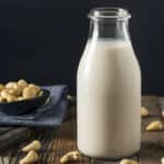 A glass bottle of cashew milk on a wooden surface with scattered cashews and a small bowl of cashews in the background.