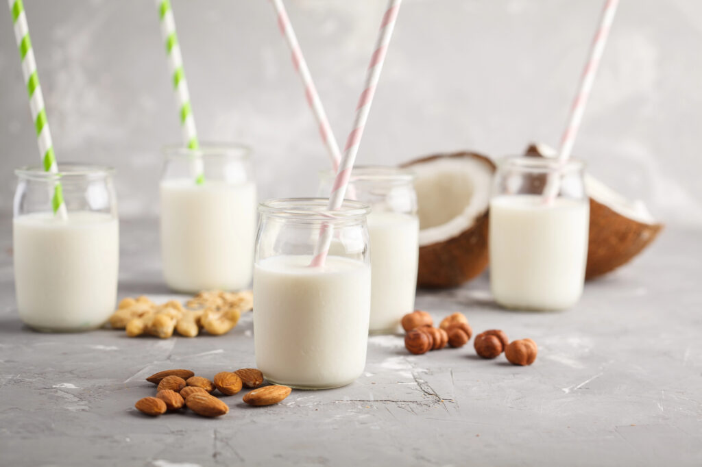 Jars of milk with straws on a gray surface, surrounded by almonds, cashews, hazelnuts, and a cracked coconut.