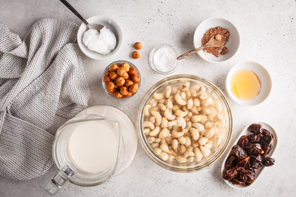 Ingredients for a recipe are arranged on a table, including soaked cashews, dates, hazelnuts, almond milk, coconut oil, cacao powder, and sweetener. A cloth lies to the side.