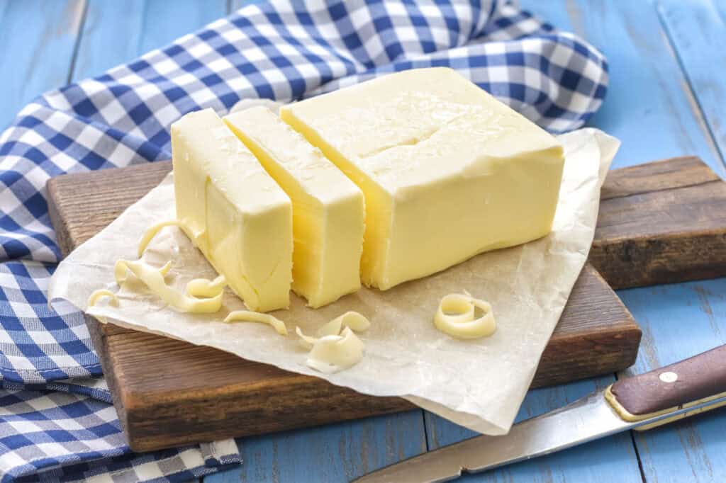 Block of butter with slices on parchment paper, placed on a wooden board. A checkered cloth is in the background, and a knife rests in the foreground.