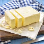 Block of butter with slices on parchment paper, placed on a wooden board. A checkered cloth is in the background, and a knife rests in the foreground.