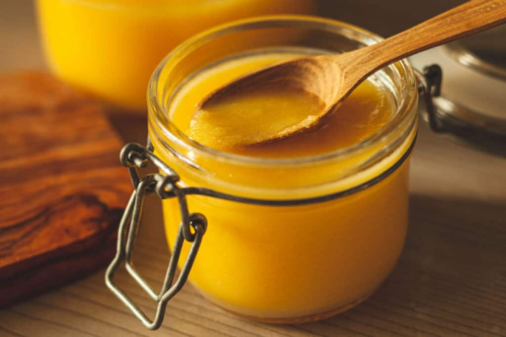 A jar of golden yellow creamy substance with a wooden spoon on top, placed on a wooden surface.