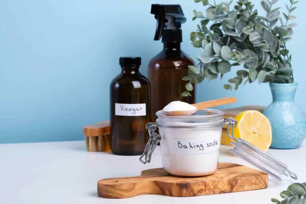 A jar of baking soda, a bottle labeled vinegar, a spray bottle, and a wooden brush on a surface with a lemon slice and eucalyptus in a blue vase.