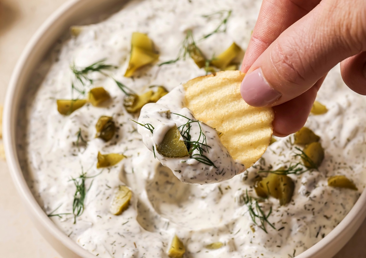 Hand dipping a ridged chip into a bowl of creamy dip with diced pickles and dill.