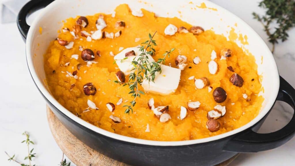 A dish filled with mashed sweet potatoes topped with chopped nuts, a pat of butter, and a sprig of thyme. The dish is placed on a wooden board, with a few thyme sprigs scattered around.
