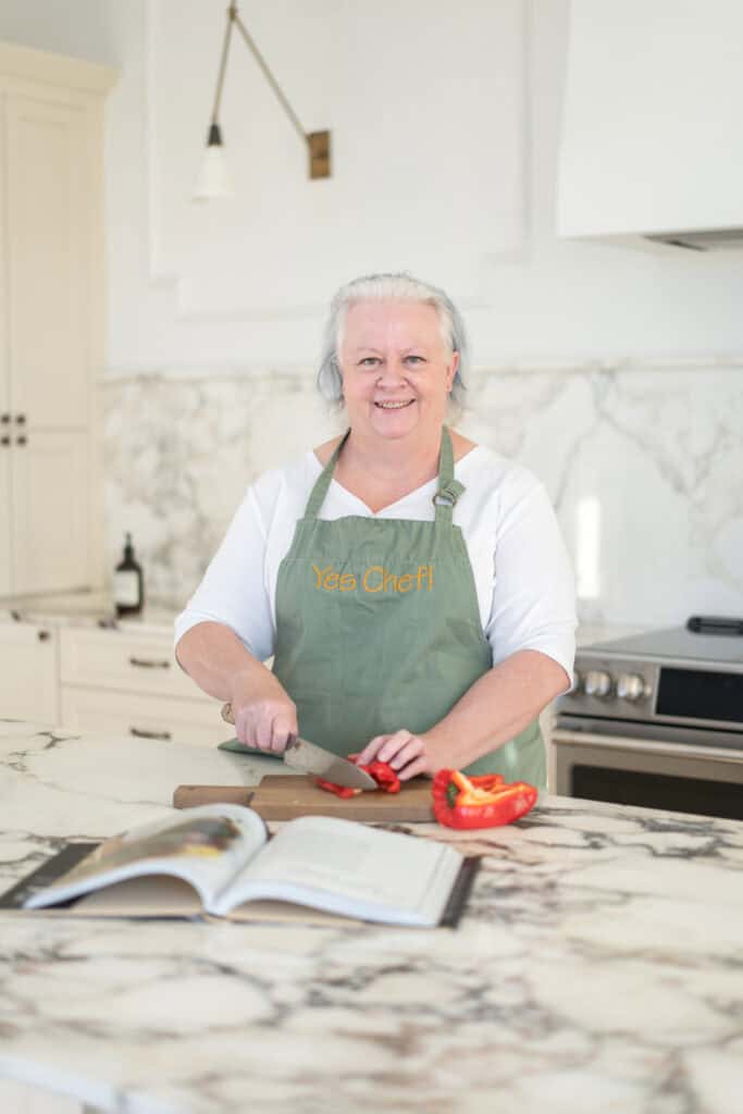 Woman in a green apron slices a red bell pepper on a marble kitchen counter with an open cookbook nearby.