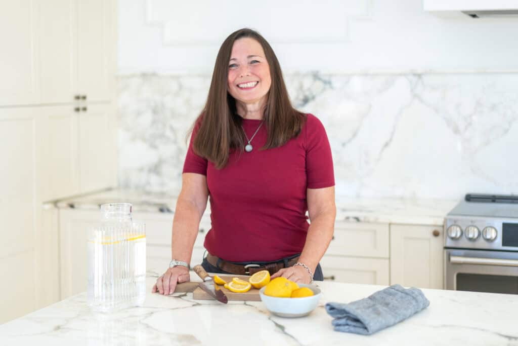 Woman in a red shirt stands in a kitchen, smiling. There are sliced oranges on a cutting board and a bowl on the counter.