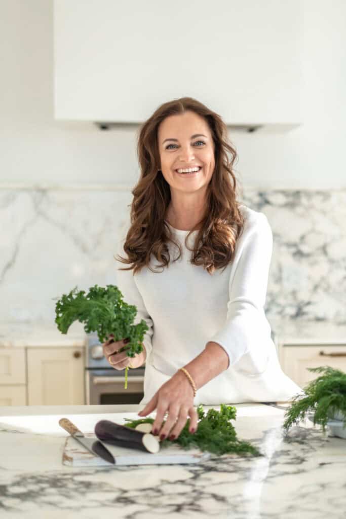 Woman in a white shirt smiles while holding leafy greens and slicing vegetables in a modern kitchen.