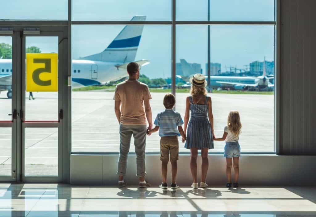 A family of four, seen from behind, holds hands while looking out at airplanes through a large airport window, embracing the joys of traveling with kids.