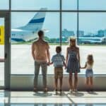 A family of four, seen from behind, holds hands while looking out at airplanes through a large airport window, embracing the joys of traveling with kids.