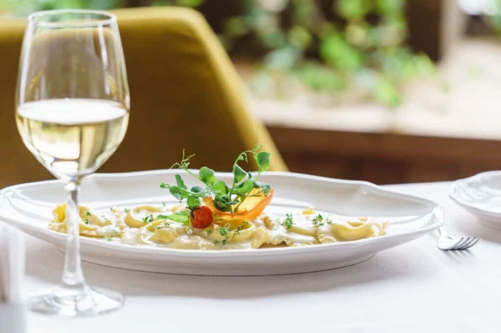 A plate of ravioli garnished with greens and cherry tomato, next to a glass of white wine, set on a white tablecloth with a blurred background.