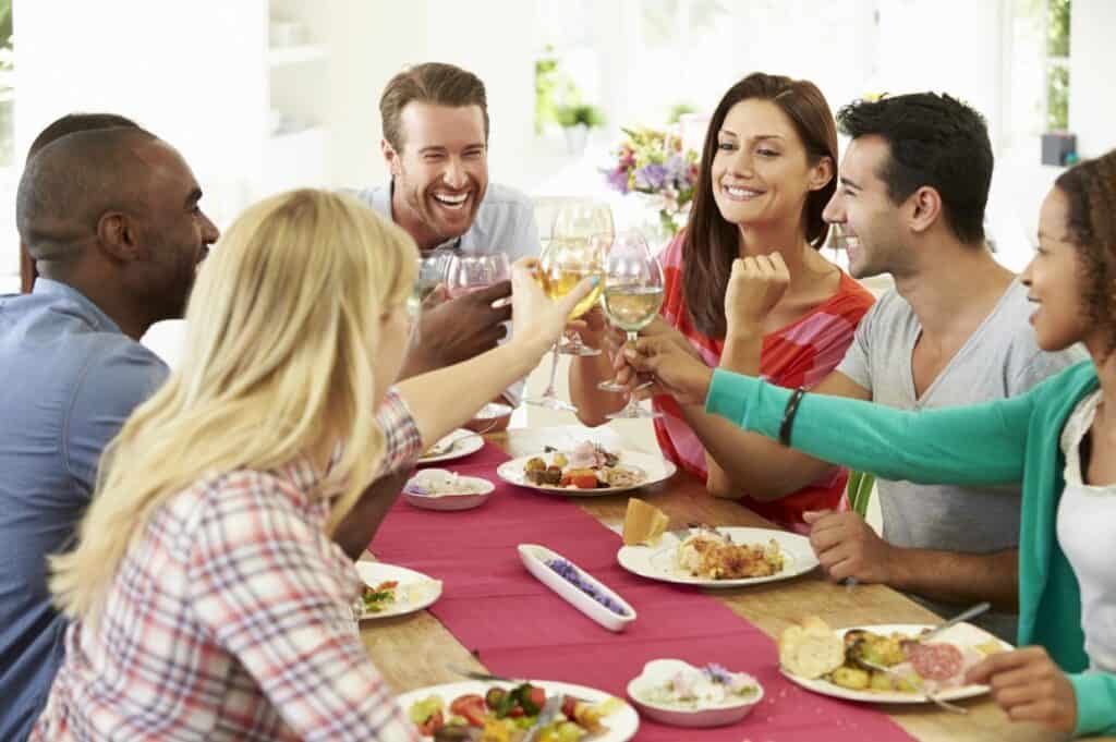 A group of six people sitting at a dining table, smiling and toasting with drinks. Plates of food are in front of them.