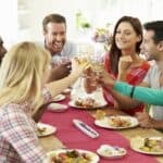 A group of six people sitting at a dining table, smiling and toasting with drinks. Plates of food are in front of them.