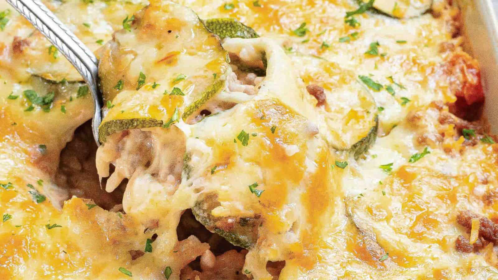 Close-up of a cheesy zucchini casserole being served with a spoon. The dish features layers of melted cheese, sliced zucchini, ground meat, and herbs, with a golden-brown crust.