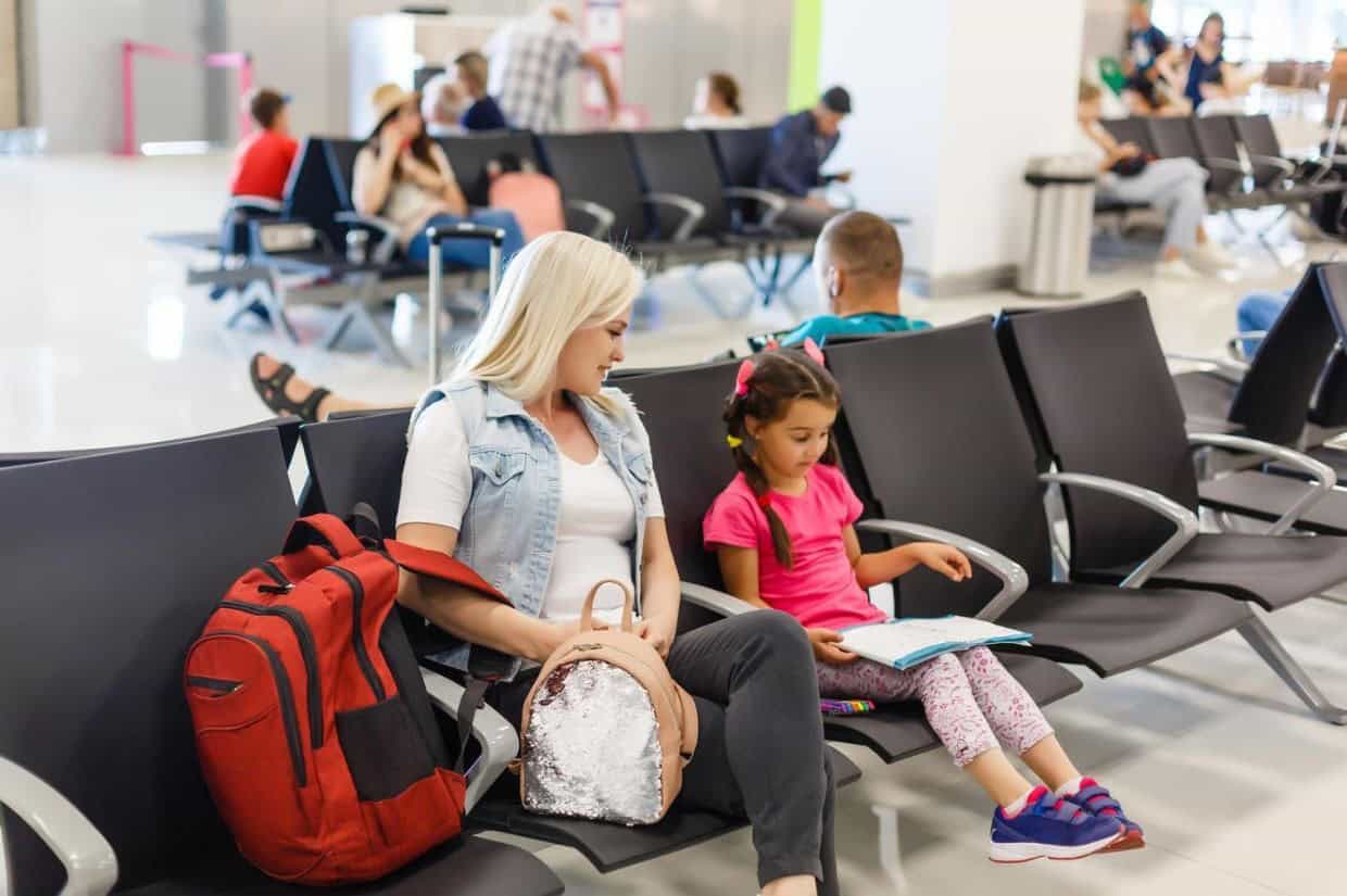 A woman and a child sit on black airport seats. The child is reading a book. Other travelers are in the background.