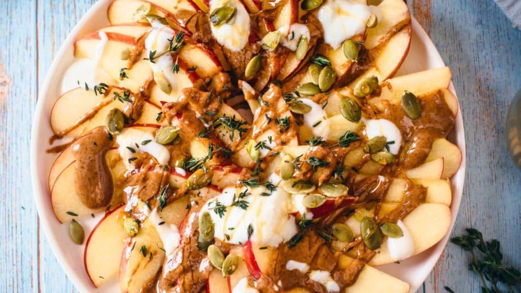 A plate of sliced apples topped with dollops of yogurt, drizzles of almond butter, sprinkled pumpkin seeds, and fresh thyme leaves, displayed on a rustic wooden surface.