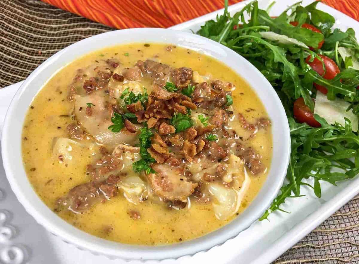 A bowl of creamy soup with herbs and meat pieces, garnished with parsley, next to a side salad with arugula, cherry tomatoes, and shaved cheese on a white plate.