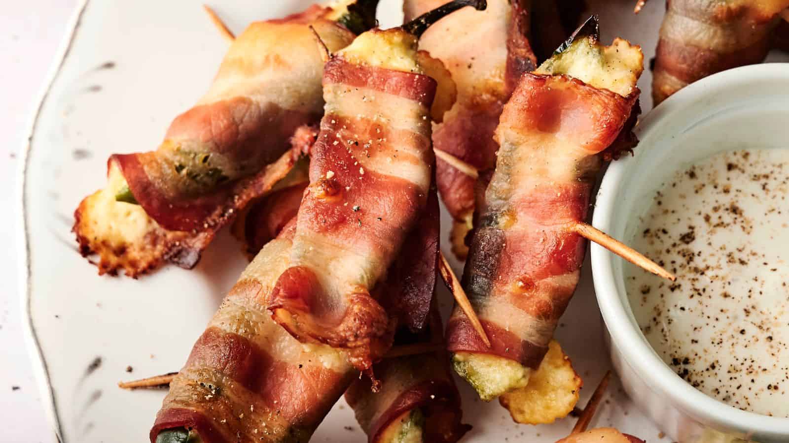 Bacon-wrapped jalape&ntilde;os filled with cheese, secured with toothpicks, on a plate next to a small bowl of dipping sauce.