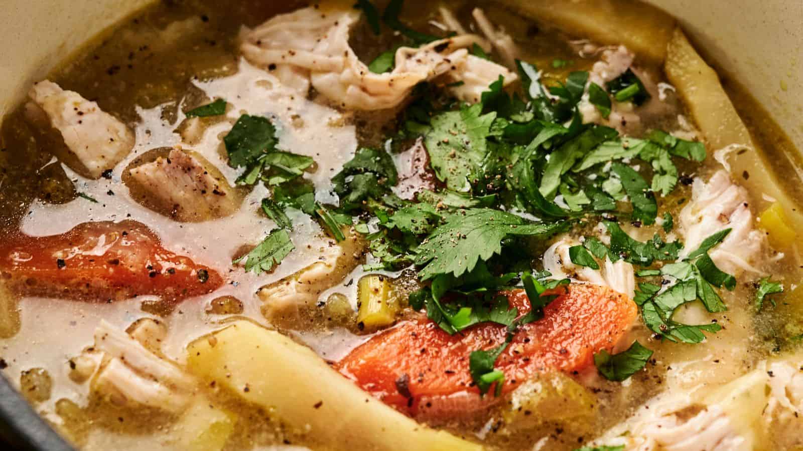 Close-up of a soup with shredded meat, tomato pieces, and slices of vegetables, garnished with chopped cilantro in a broth.