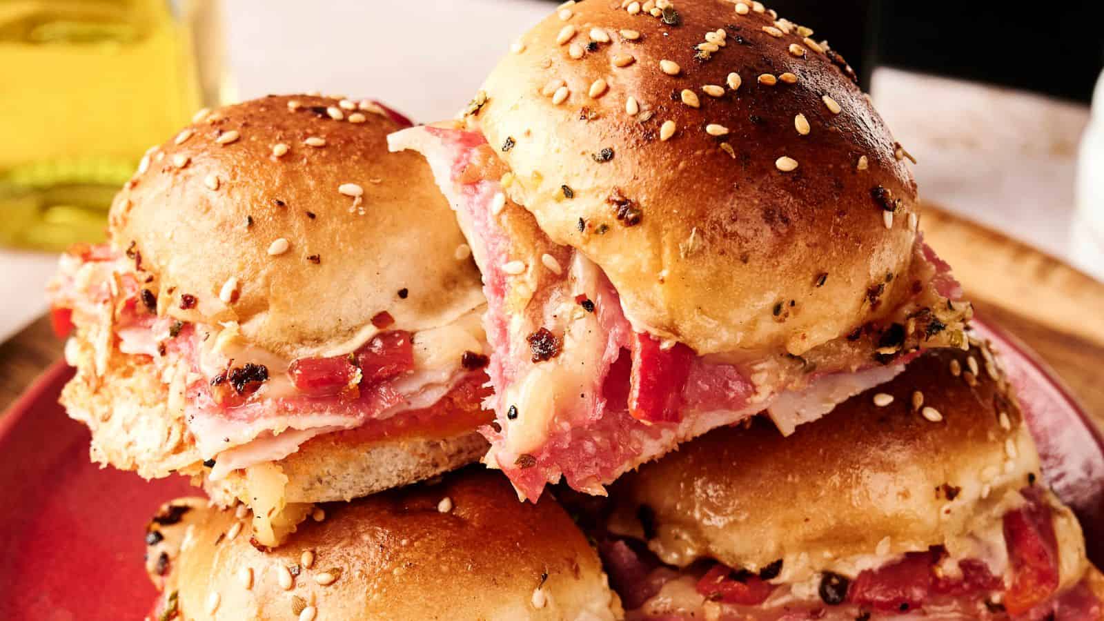 A close-up of three seasoned, sesame-seed-topped slider sandwiches filled with layers of sliced meat and cheese on a red plate.