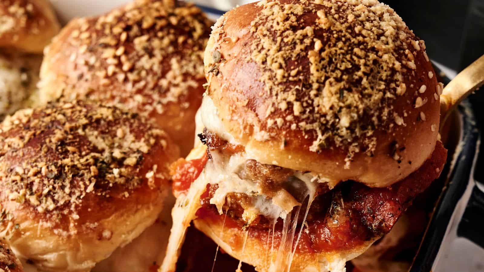Close-up of cheese-stuffed sliders topped with herbs and crumbs, with one slider being lifted to reveal melted cheese stretching from the tray.