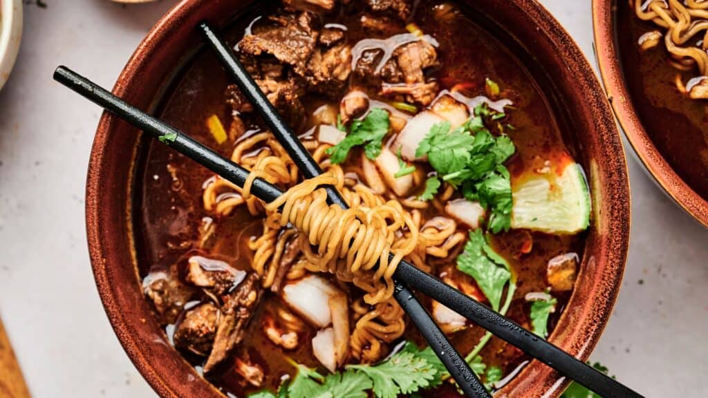 A bowl of noodle soup with beef pieces, chopped onions, and cilantro, garnished with lime. Black chopsticks rest across the bowl with noodles wrapped around them.