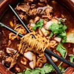 A bowl of noodle soup with beef pieces, chopped onions, and cilantro, garnished with lime. Black chopsticks rest across the bowl with noodles wrapped around them.