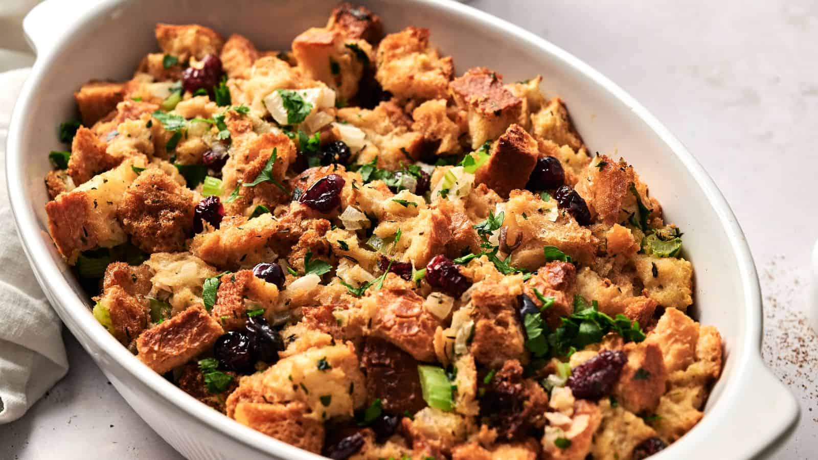 A close-up of a casserole dish filled with stuffing made of bread cubes, cranberries, chopped herbs, and celery.