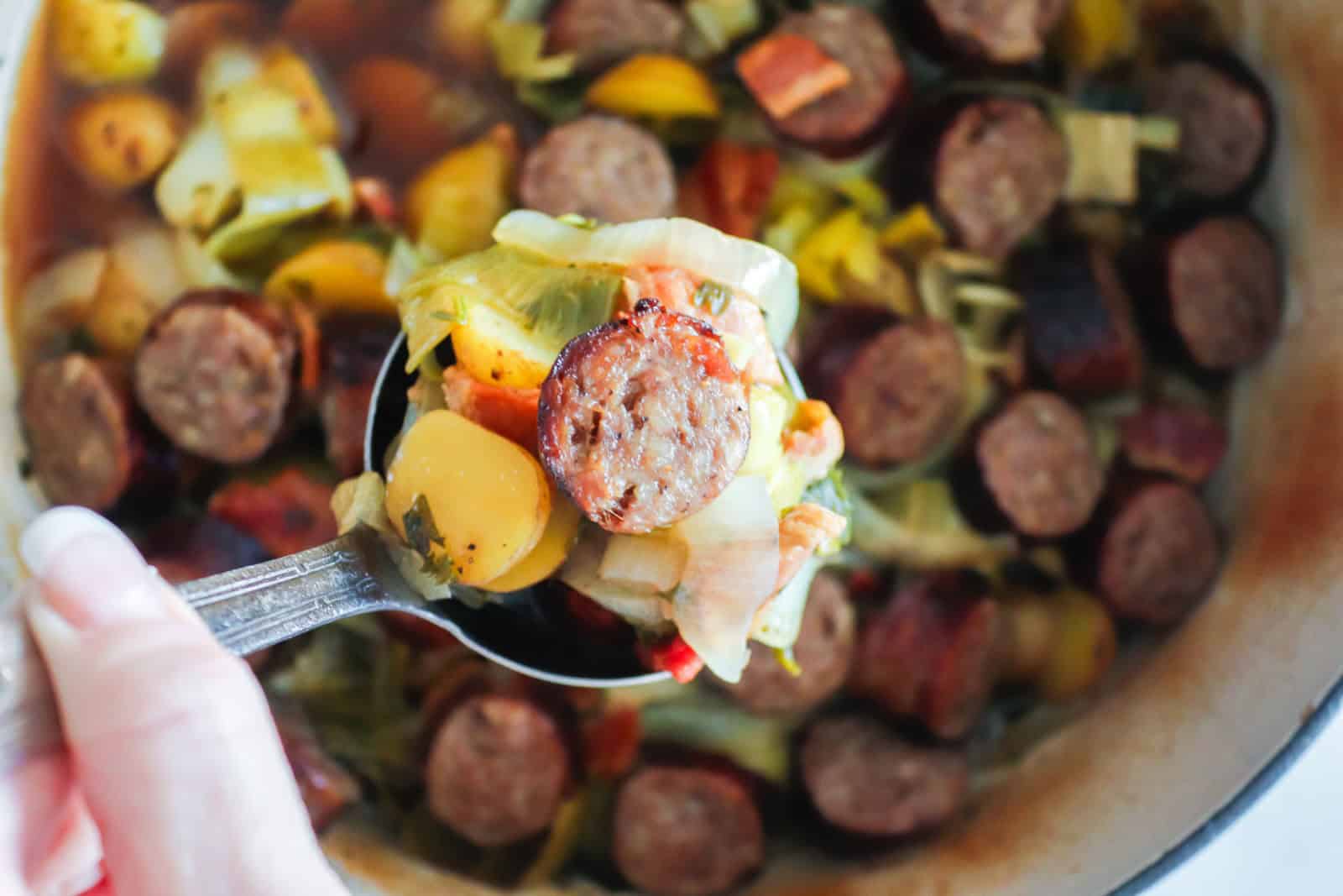 Sliced sausages with cooked onions, potatoes, and vegetables in a white baking dish.