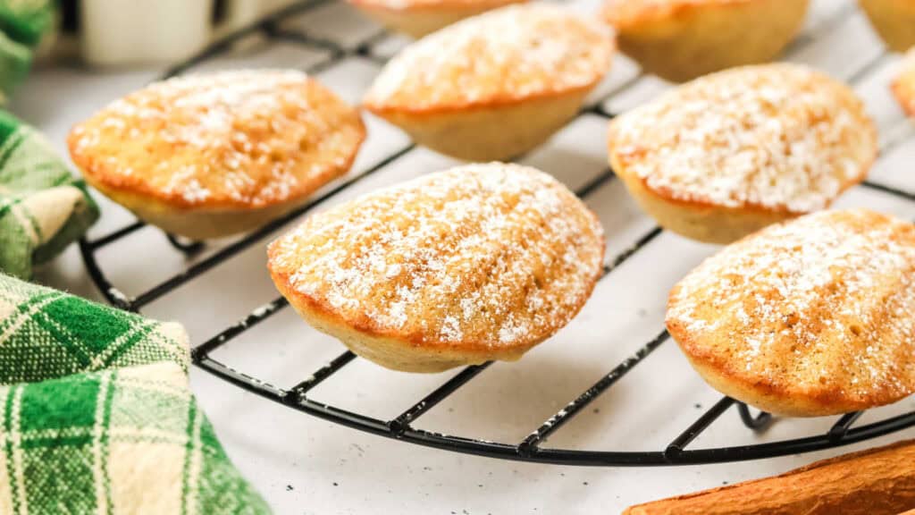 Madeleines dusted with powdered sugar on a cooling rack, next to a green plaid cloth.