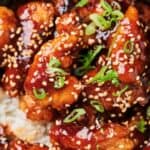 Close-up of glazed chicken pieces topped with sesame seeds and chopped green onions, served over a bed of white rice.