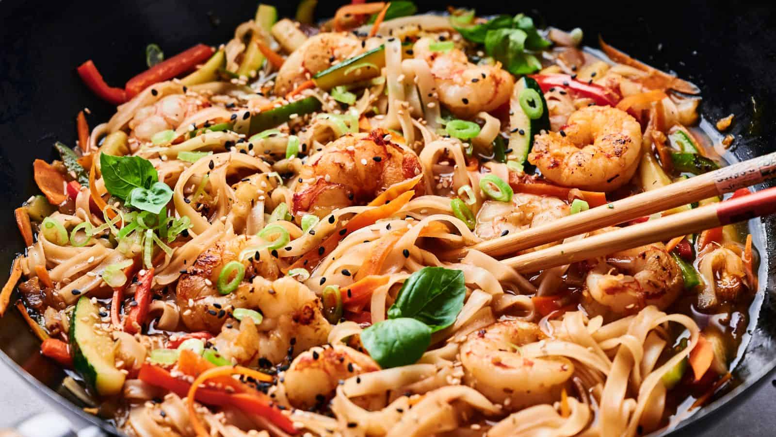 A colorful dish of shrimp stir-fry with noodles, vegetables, and sesame seeds, garnished with basil leaves and green onions. Chopsticks rest on the dish.