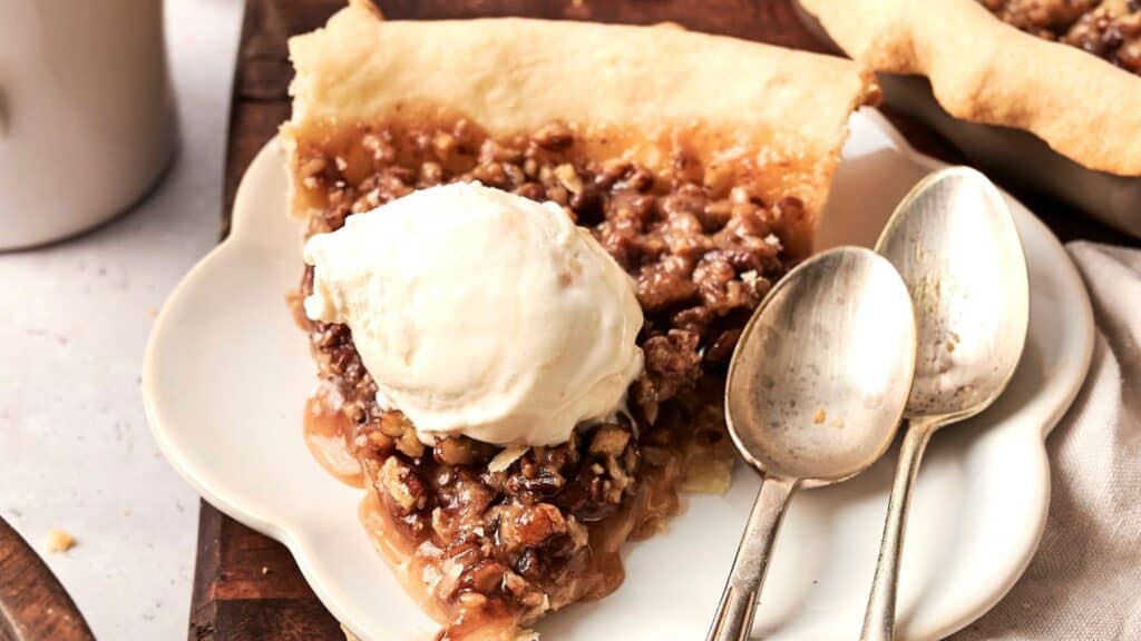 A slice of pecan pie topped with a scoop of vanilla ice cream on a plate, accompanied by two spoons.
