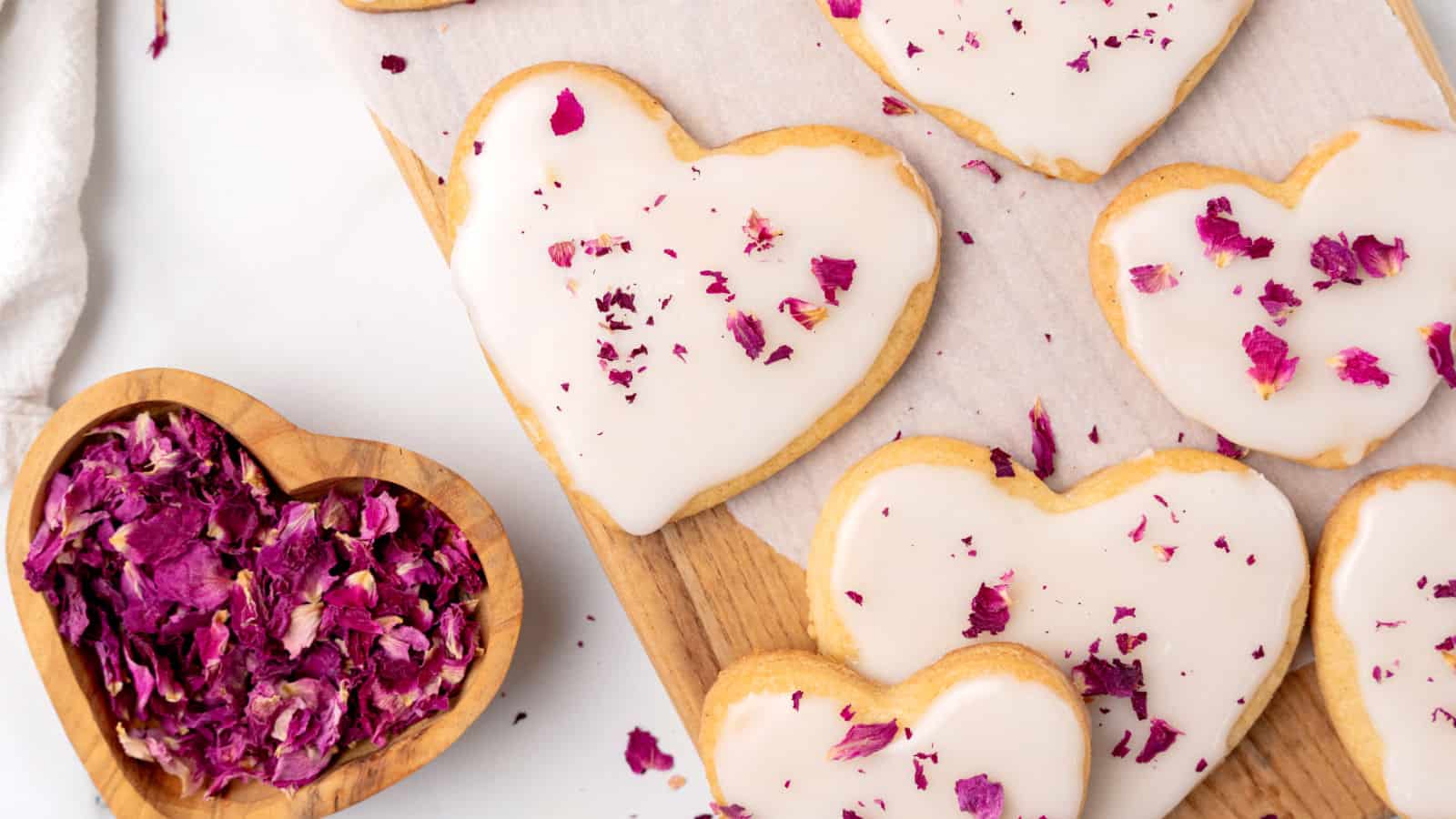 Heart-shaped cookies with white icing and rose petal decorations on a wooden board, next to a heart-shaped bowl filled with rose petals.