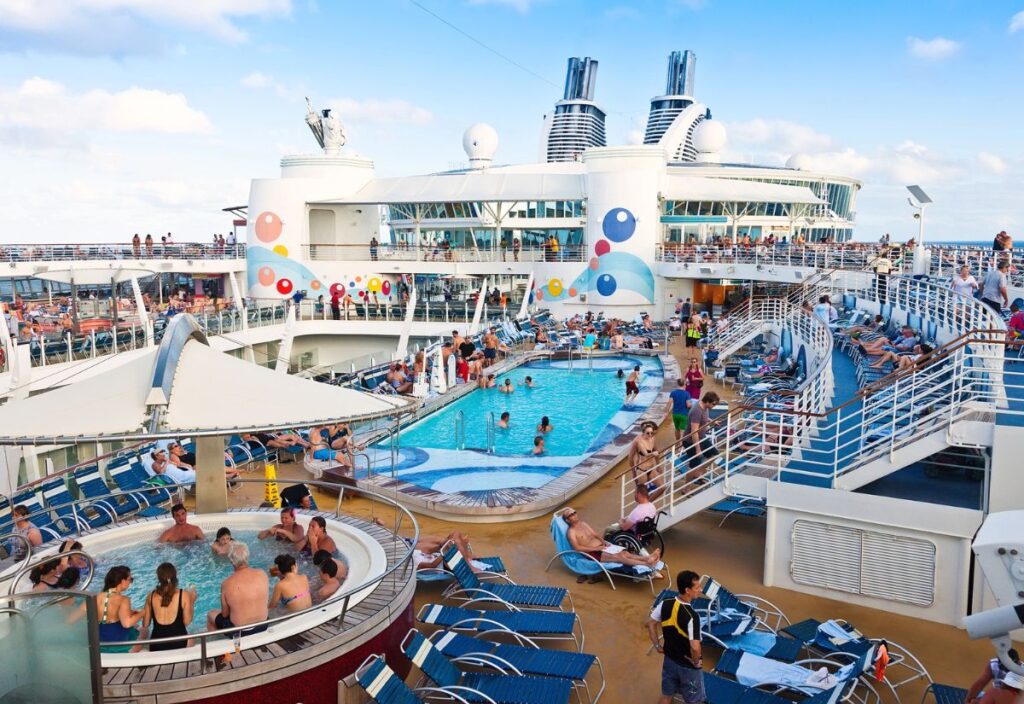 People relax by a large pool on a cruise ship deck, with loungers and a hot tub, under a clear sky.